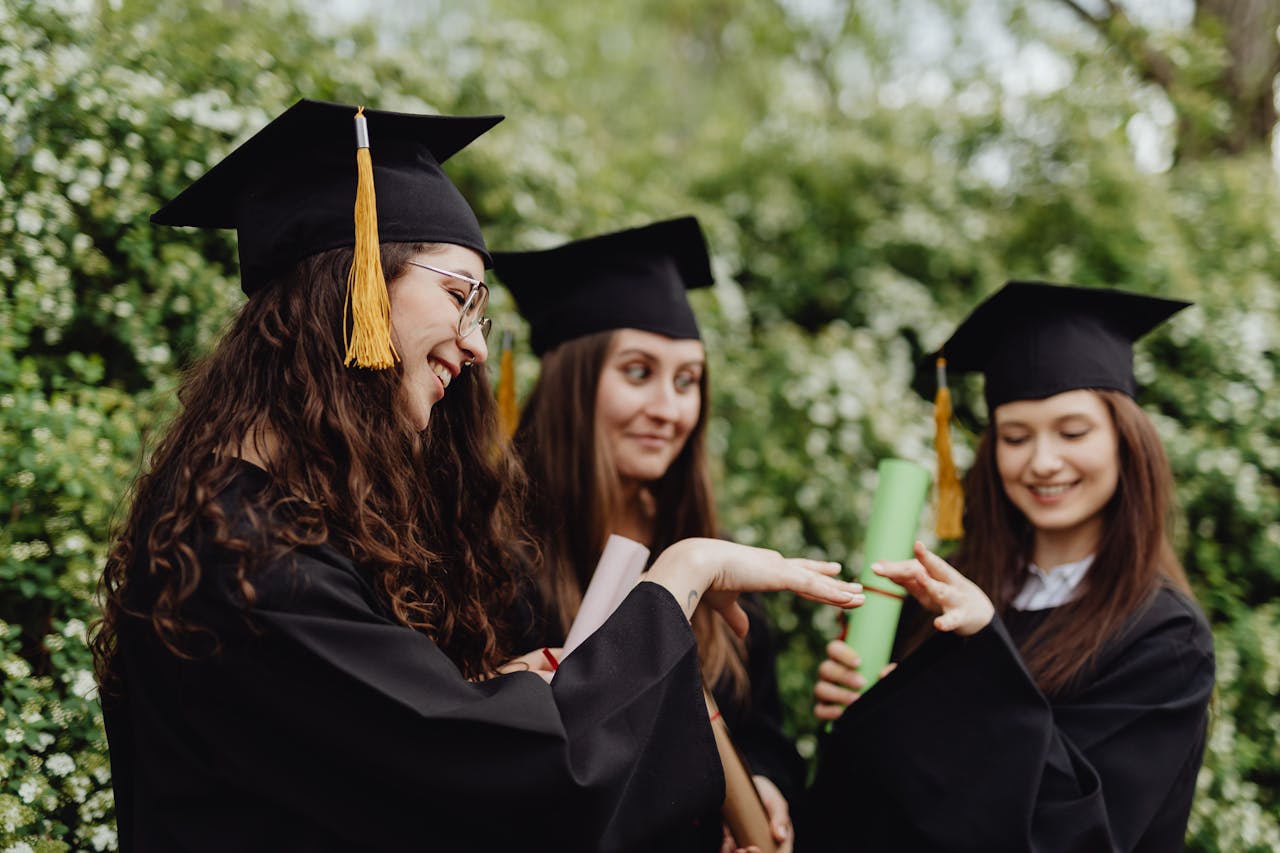 Three young women celebrate graduation outdoors with caps and gowns, showcasing their diplomas and happiness.